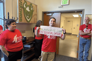 people in red shirts holding sign. Sign “Recognize language equity. Pay bilingual employees.” 